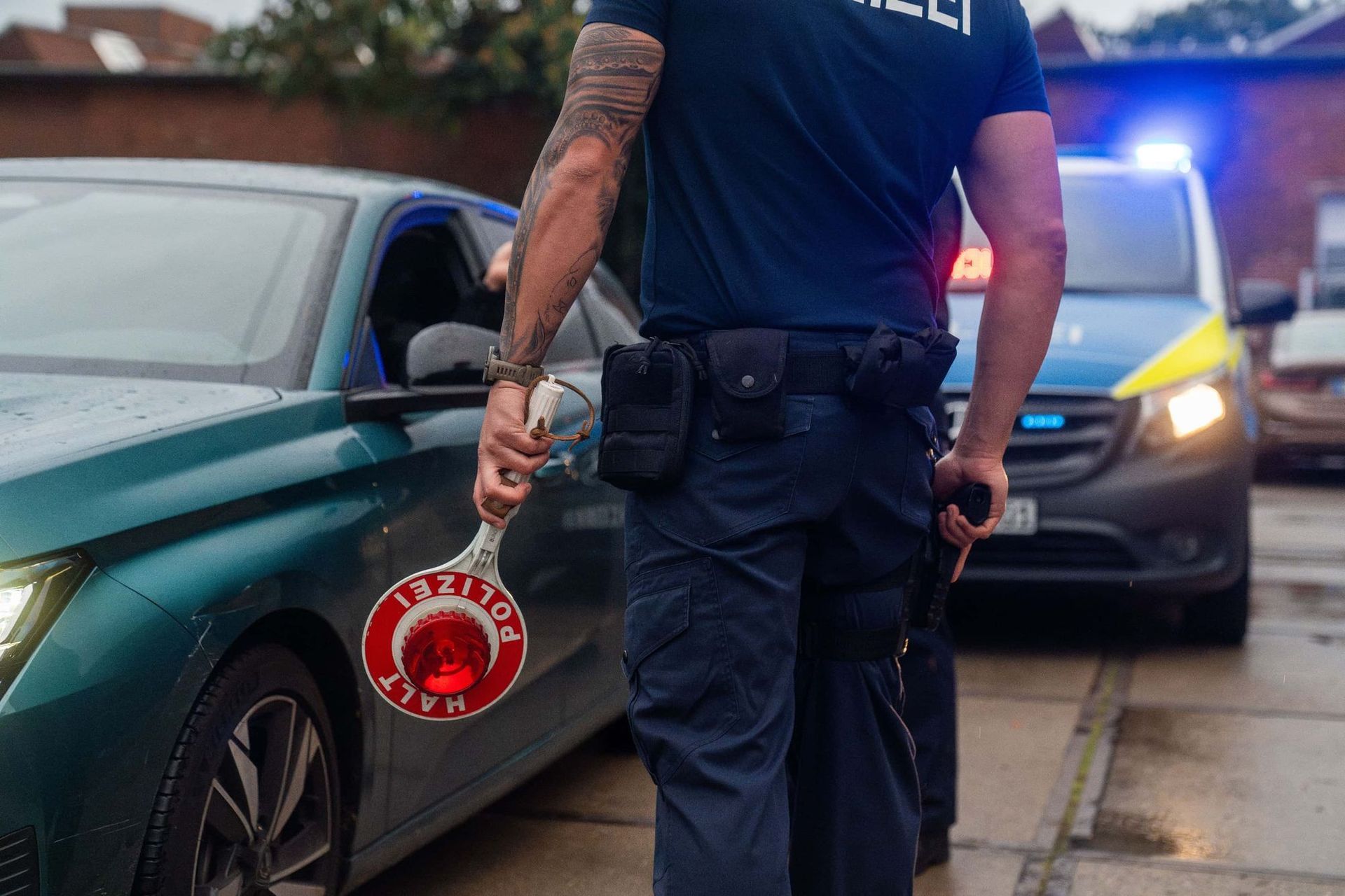 Polizist mit Stoppschild nähert sich einem grünen Auto; im Hintergrund ein Polizeiauto mit Blaulicht.