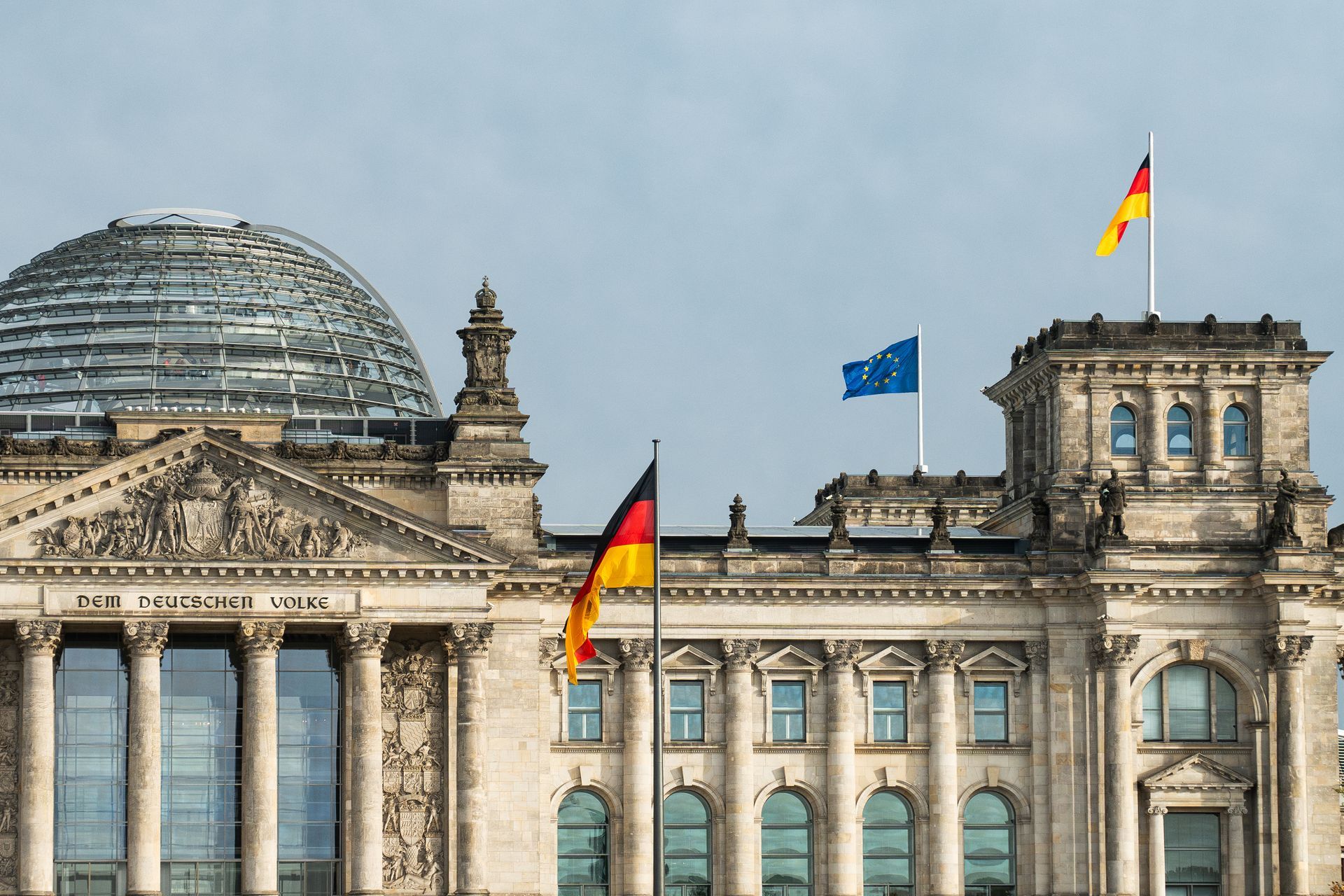 Reichstagsgebäude in Berlin mit Deutschland- und EU-Flaggen vor bewölktem Himmel.